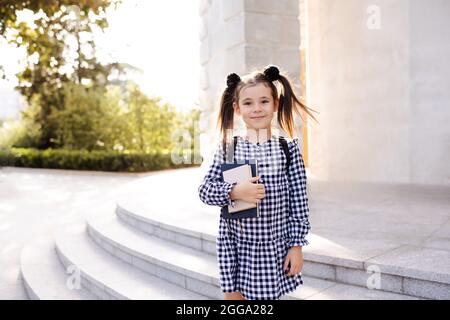 Sweet smiling young child girl 5-6 year old wear checkered black and white dress and backpack holding books stand outdoors close up. Looking at camera Stock Photo