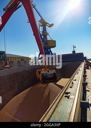 a crane with a manipulator bucket loads wheat onto a dry cargo ship at the berth in the port. Loading and unloading of bulk cargo for export by sea. S Stock Photo