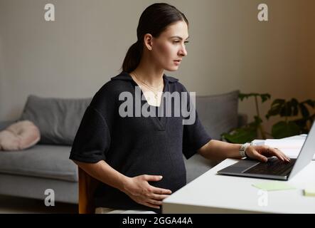 Busy pregnant young Caucasian woman sitting at table in living room and using laptop while working remotely Stock Photo