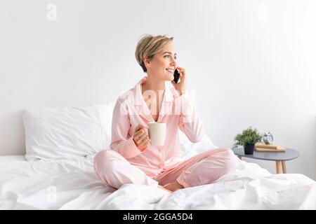 Happy woman sitting on bed talking on phone drinking coffee Stock Photo