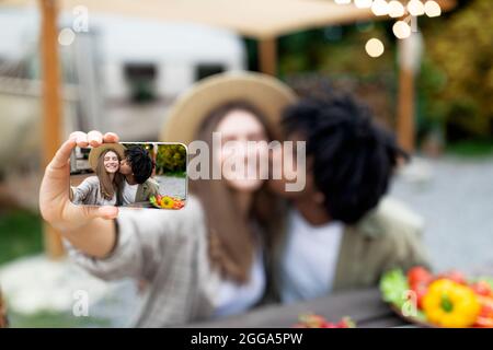 Affectionate Afro guy kissing his girlfriend, taking selfie on mobile phone near motorhome on camping trip in autumn Stock Photo