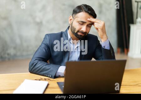 Upset Indian businessman in formal suit has strong headache, sitting in front of laptop and holding forehead, feels tired and exhausted, sad eastern man worrying, feels anxiety, suffers from migraine Stock Photo