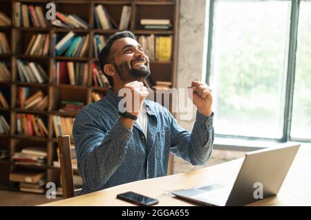 Excited surprised middle eastern businessman got unbelievable notification about win, mixed-race indian guy screaming yes, raising fists up in triumph gesture, sits at workplace in loft office Stock Photo