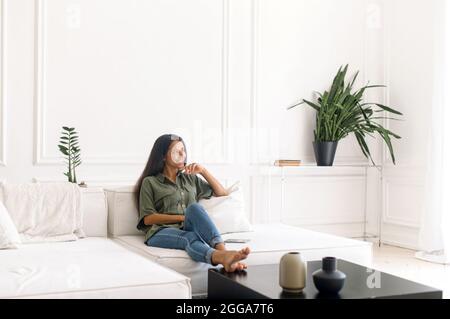Carefree and calm attractive young indian woman sitting on the comfortable white couch in cozy apartment in relaxed atmosphere, smiling mixed-race lady enjoys lazy weekend at home, daydreaming alone Stock Photo