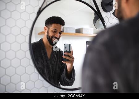 Young man with bathrobe indoors in bathroom at home, taking selfie. Stock Photo