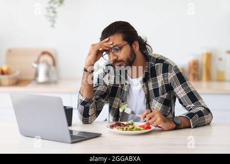 Exhausted middle-eastern man freelancer having snack while working from home Stock Photo
