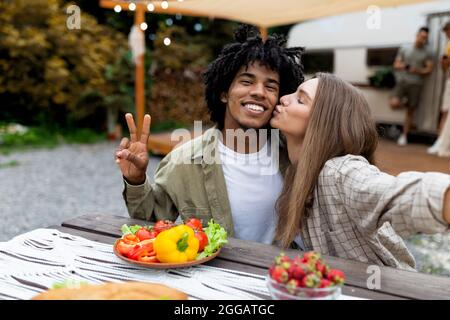 Young multinational couple making selfie during lunch, kissing, showing peace gesture near camper van Stock Photo