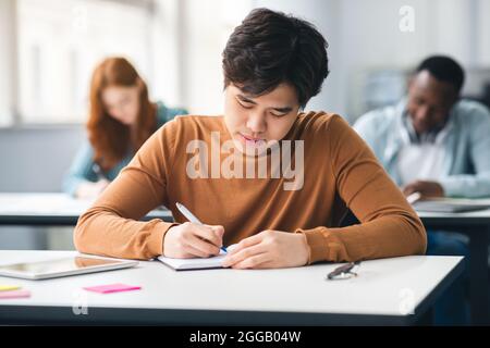 Asian student sitting at desk in classroom writing in notebook Stock Photo