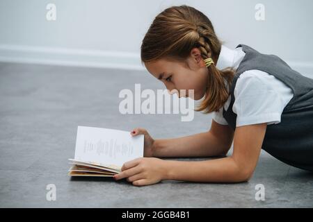 Side view of a little schoolgirl in a uniform lying on the floor, reading a textbook Stock Photo