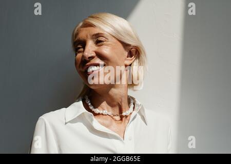 Minimal portrait of elegant mature woman lit by sunlight against white wall and smiling, copy space Stock Photo