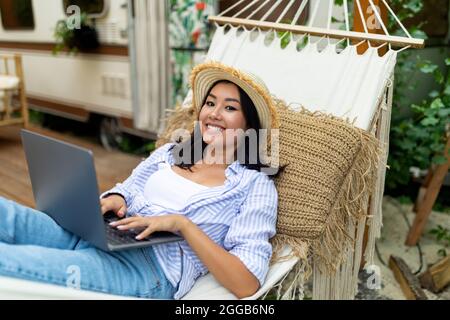 Joyful Asian lady using laptop computer while lying in hammock near RV on autumn camping trip Stock Photo