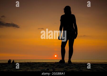 Rear view silhouette of an isolated young woman, in sports cap, looking out to sea at sunset on a UK beach, staycation summer holiday. Stock Photo