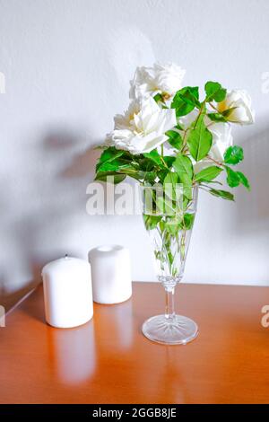 White roses in vase, white candles on wooden commode across white wall. Interior design. Home decor. Stock Photo