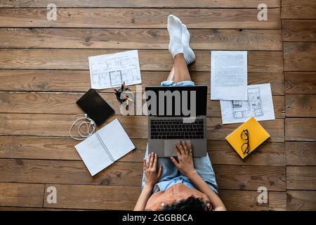 Top view of young lady with laptop, study or work materials and tablet pc sitting on wooden floor indoors, mockup Stock Photo