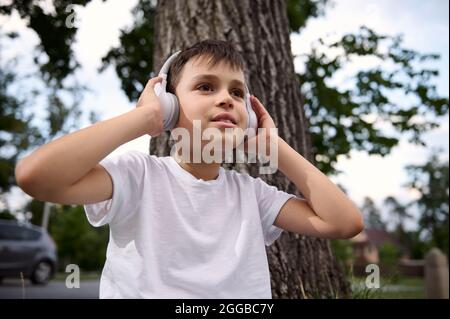 Portrait of handsome adorable child boy, wearing wireless headphones and enjoying listening to music, resting during recreation between classes in the Stock Photo