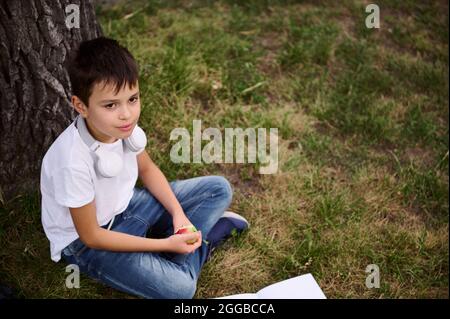 Handsome school child boy sitting on green grass of public park , wearing headphones around his neck, looking away and holding an apple in his hand. P Stock Photo