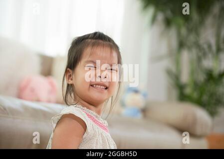 Cheerful pretty little girl smiling and squinting eyes when standing in living room of apartment Stock Photo