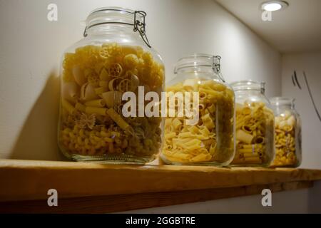 Four glass jars standing on the shelf and filled with various types of pasta. Stock Photo