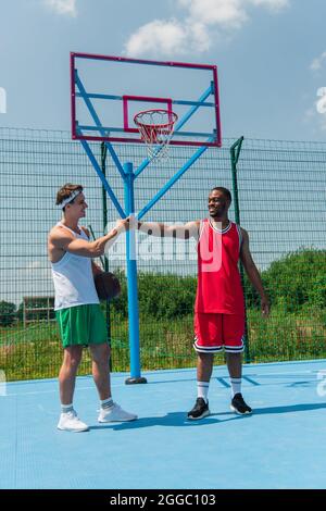 Positive sportsman with basketball ball doing fist bump with african american friend on playground Stock Photo