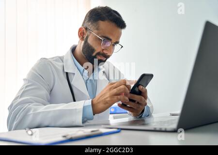 Indian serious male doctor in clinic uses cell mobile apps and laptop computer. Stock Photo