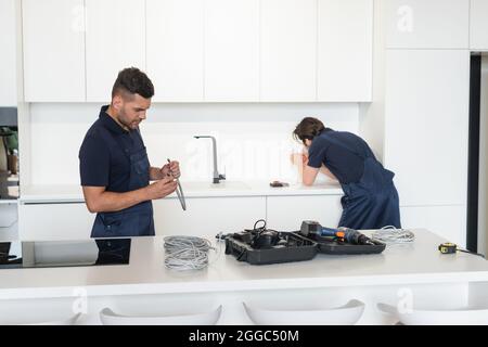 workman holding metal hose near electrician working with power sockets in kitchen Stock Photo