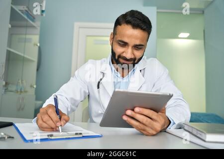 Indian happy male doctor in clinic using tablet device writing health data. Stock Photo