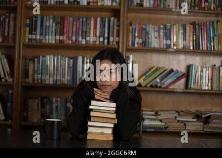 Shot of an Asian girl with her head held on hands on the books in the library and looking forward. Stock Photo
