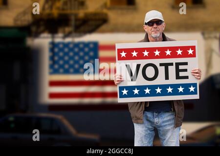 Man with cap, blue jeans and sunglasses holding a cardboard sign text VOTE with american stars and stripes flag on a wall in the background. American Stock Photo