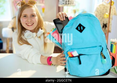 smiling modern pupil with tablet PC, workbooks and blue backpack in white sweatshirt packing for school at home in sunny day. Stock Photo