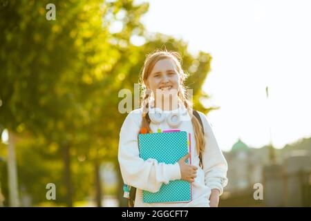 Portrait of happy modern child in white sweatshirt with workbook and headphones going to school outdoors in the city. Stock Photo