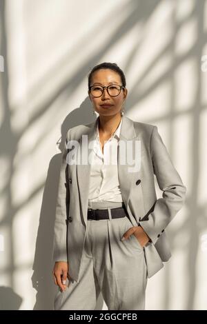 Young confident businesswoman of Asian ethnicity standing in front of camera against white wall Stock Photo