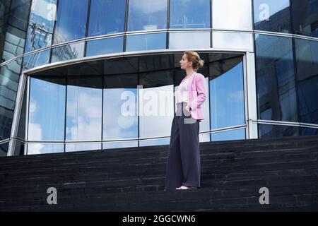Young blonde of millennium with strict hairstyle in pink elegant clothes stands on steps near modern office building in full growth, half-turned to ca Stock Photo