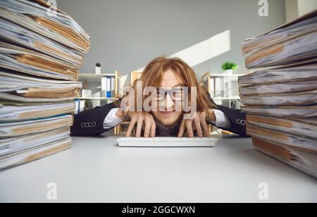 Happy office worker sitting at desk with stacks of papers and typing on wireless keyboard Stock Photo