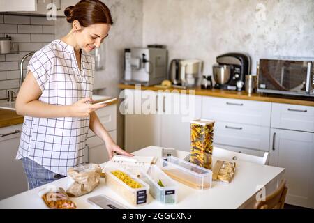 Smiling young beautiful business female taking writes a list of products to buy in paper diary at modern Nordic style kitchen Stock Photo