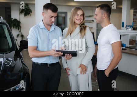 Salesperson talking with couple and help choosing new car. Man and woman in car showroom. Man using tablet to shop opportunity of different models Stock Photo