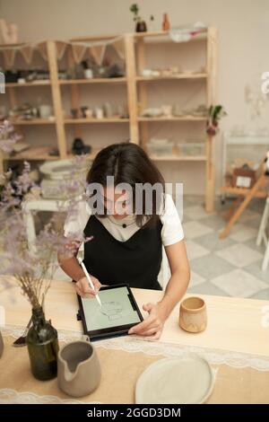 High angle of creative young female ceramics designer drawing sketch of new clayware on tablet with stylus pen while working in studio Stock Photo