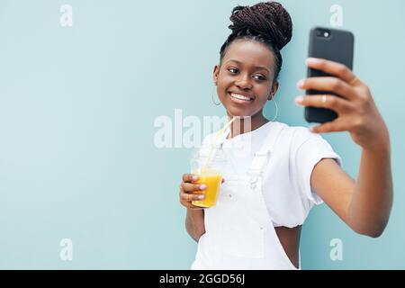 Young smiling woman holding a juice and taking a selfie while standing outdoors at blue wall Stock Photo