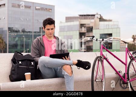 young student sitting outdoors using tablet next to his bicycle, concept of technology and urban lifestyle Stock Photo