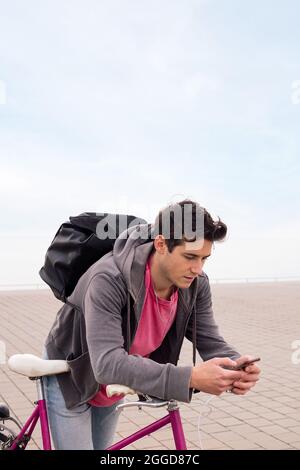 vertical photo of a handsome young student with backpack leaning on a bike using a mobile phone, concept of communication and ecological urban transpo Stock Photo