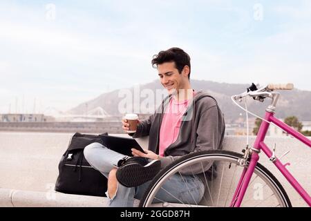 happy young man sitting outdoors with coffee smiling and watching video on a tablet next to his bike, concept of technology and urban lifestyle, copy Stock Photo
