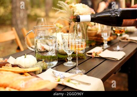 Glass of orange wine placed near dishware and bouquet of fresh flowers amidst assorted fruits on wooden table on autumn day in garden. Season party or Stock Photo