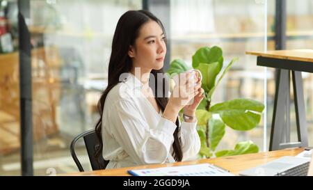 Thoughtful asian businesswoman, positive pensive thinking, looking away, drinking coffee Stock Photo