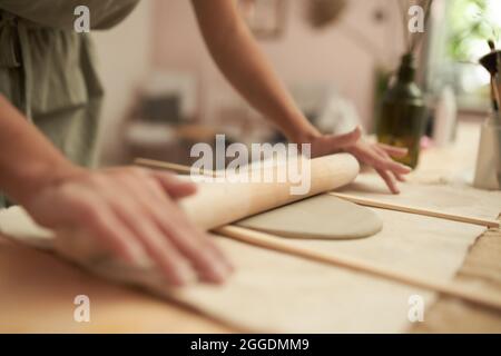 Crop anonymous craftswoman using rolling pin and wooden sticks while making slab of clay for handmade pottery in workshop Stock Photo