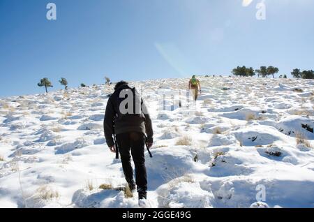 two men with backpacks going up a large slope towards the peak of a snowy mountain Stock Photo