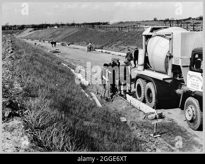 A view of the construction of the Birmingham to Preston Motorway (M6), looking north along a slip road on the east side of Section D of the motorway and showing a group of men in the foreground, standing beside a concrete mixer lorry. The work on the Birmingham to Preston Motorway (M6), between junctions J13 to J16 started in June 1960 and was carried out by John Laing Construction Ltd. Section D was 7 1/2 miles in length between Keele and Barthomley. Stock Photo