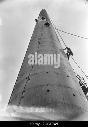 Construction workers suspended in harnesses working on the surface of a chimney stack at the South Eastern Gas Board's (SEGAS) Power Station on the Isle of Grain. In the late 1950's, the South Eastern Gas Board (SEGAS) set up a power plant on the Isle of Grain. The plant was adjacent to the Grain refinery, and was designed to produce gas from petroleum products. In the negative register, 'Lytag S. E. Gas Board, Isle of Grain, Kent' is recorded next to this image. Lytag is a lightweight aggregate material that can withstand high temperatures. According to the April 1963 edition of Laing's month Stock Photo