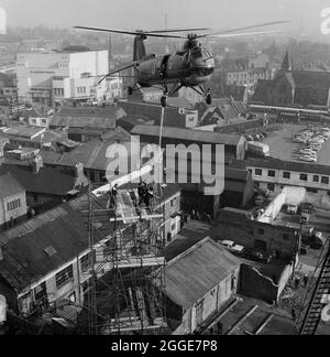 A view looking north from the roof of Coventry Cathedral, showing a RAF Belvedere helicopter attempting to hoist the 80ft bronze spire for the new cathedral from a scaffolding tower on site, with the city of Coventry in the background including the bus station and The Coventry Theatre. The photograph was taken during 'Operation Rich Man' a joint project involving Royal Air Force staff and Laing staff. This involved a RAF Belvedere helicopter hoisting in to place the 80ft bronze spire on to the new cathedral. The part of the operation to lower the 1/2 ton cross that sits on top of the spire had