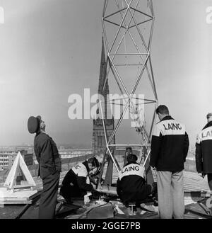 Laing workers on the roof of Coventry Cathedral attaching the 80ft bronze spire to a 1 yard square plate, while a RAF officer looks up at a RAF Belvedere helicopter (out of view). The photograph was taken during 'Operation Rich Man' a joint project involving Royal Air Force staff and Laing staff. This involved a RAF Belvedere helicopter hoisting in to place the 80ft bronze spire on to the new cathedral. The part of the operation to lower the 1/2 ton cross that sits on top of the spire had to be postponed due to windy conditions. Stock Photo