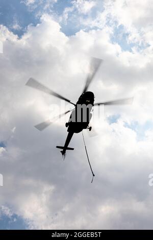 Silhouetted flying helicopter with blurry moving propeller and carrying sling on overcast sky. Stock Photo