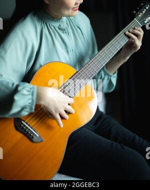 Smiling young asian woman practices to play wooden solid spruce acoustic guitar in dark room. Stock Photo